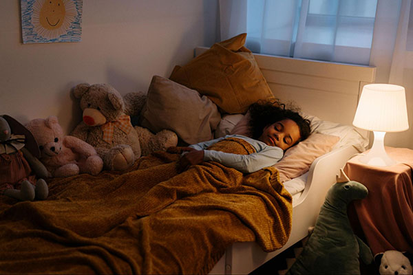 Enfant s'endormant paisiblement dans son lit, entouré de peluches, avec une lampe de chevet allumée créant une ambiance douce et rassurante