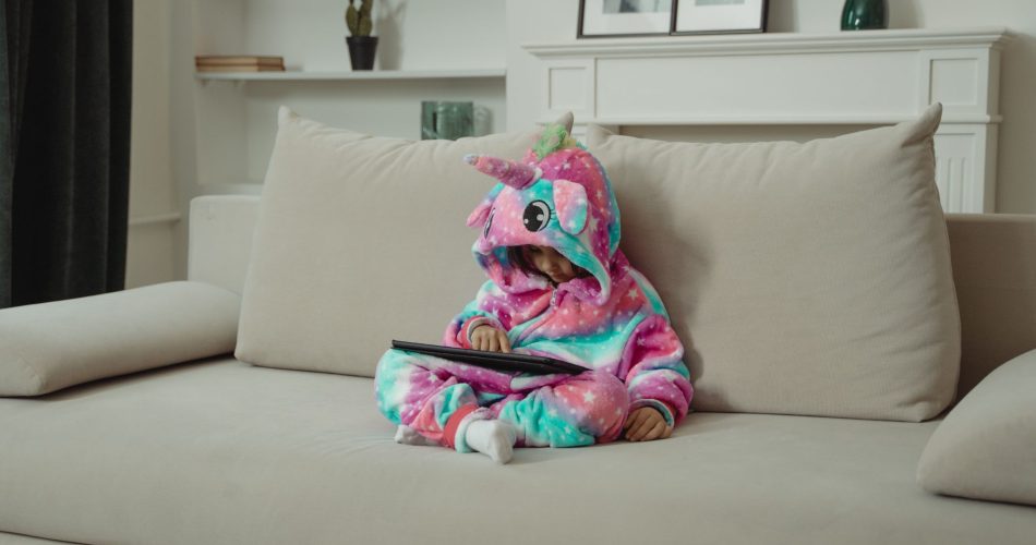 Enfant en pyjama licorne utilisant une tablette, assis sur un canapé dans un salon.
