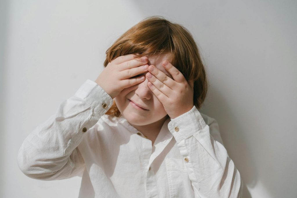 Enfant se couvrant les yeux avec les mains devant un mur clair.