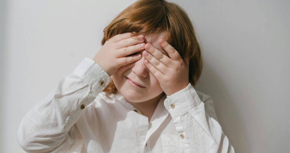 Enfant se couvrant les yeux avec ses mains devant un mur clair.