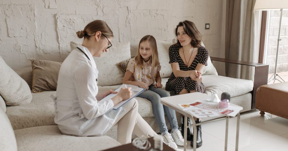Une femme en blouse blanche échange avec une mère et sa fille assises sur un canapé dans un salon lumineux.