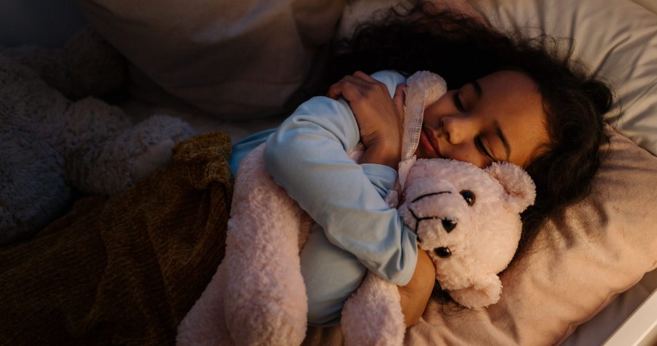 Jeune enfant endormie dans son lit, serrant un grand ours en peluche contre elle sous une lumière douce et chaleureuse.