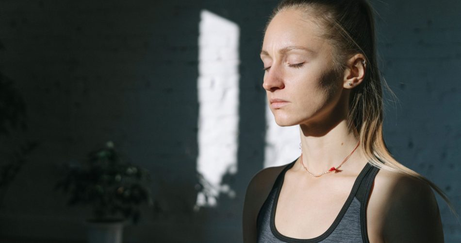 Femme aux yeux fermés, vêtue d’un débardeur de sport gris, éclairée par la lumière du soleil dans une pièce sombre.
